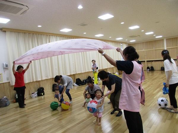 二人の先生がシーツを広げ持ち上げてトンネルを作りその下をボールを持ったお母さんと子供たちが通って遊んでいる写真