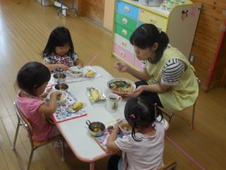3人の女児と1名の1日保育士体験している保護者が同じテーブルに座って給食を食べている様子の写真