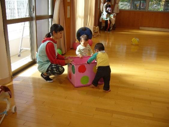 ピンク色の箱につかまり立ちをしている2人の乳児とそれを見守っている一日保育士体験のお母さんの写真
