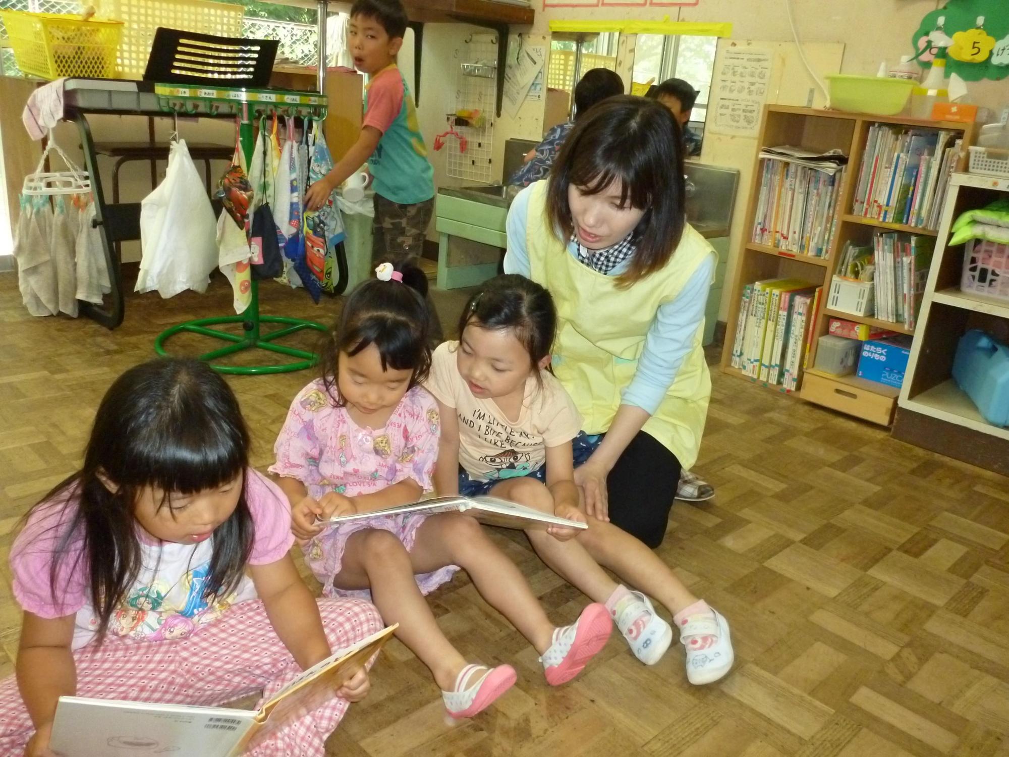 保育園の教室の中で一日保育士体験のお母さんと三人の女児が絵本を読んでいる後ろで男児がタオルで手を拭いている写真