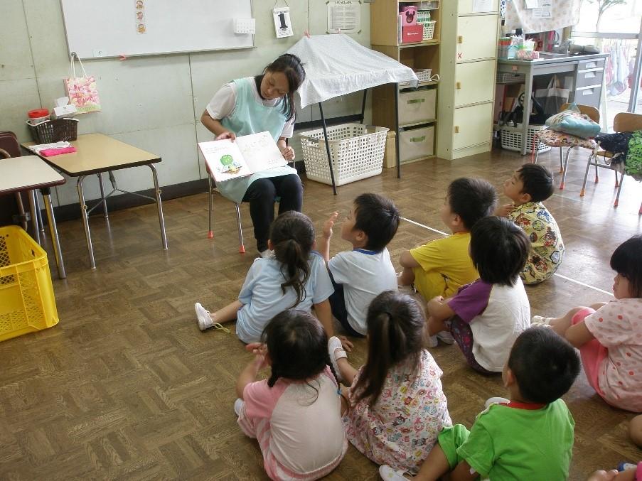 保育園の教室で椅子に座って絵本を読んでいる一日保育士体験のお母さんとお話を聞いている9人の園児たちの写真