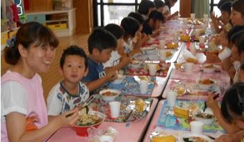 1列にテーブルを並べて向かい合わせに座りみんなで一緒に給食を食べている写真