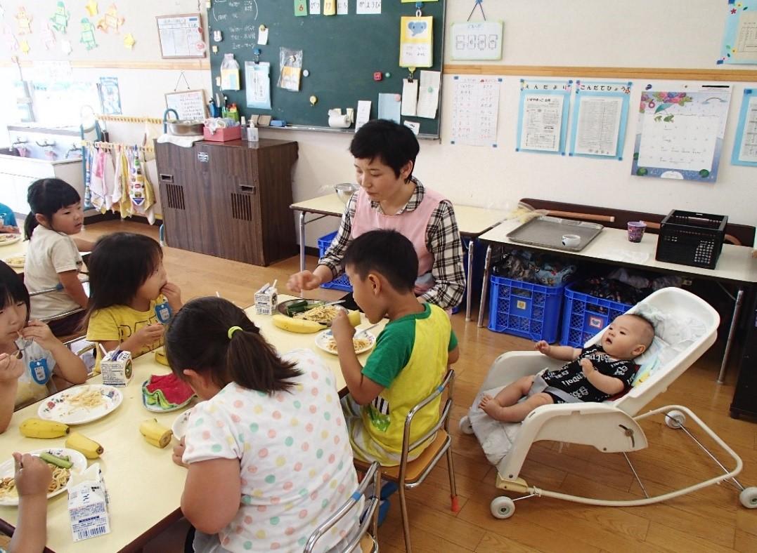 園児5人と同じテーブルを囲んで給食を食べている保護者とその横にベビーラックに入っている赤ちゃんの写真