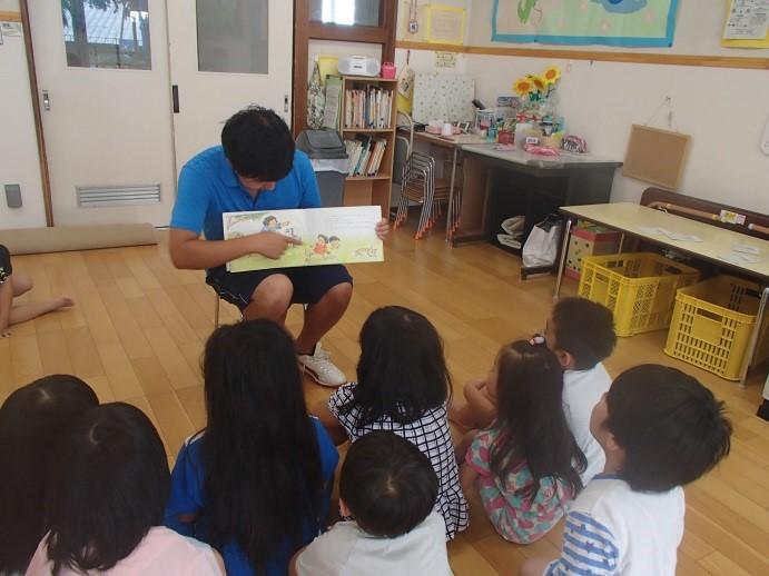 子供たちの前に椅子に座り絵本の絵を指さしながら読んでいる男性保護者の写真