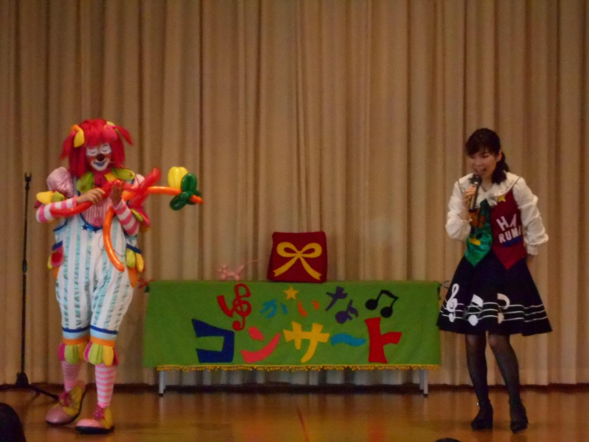 ステージ右側にマイクを持った歌のお姉さんが立ち、左側にバルーンアートで何かを作っているピエロの写真