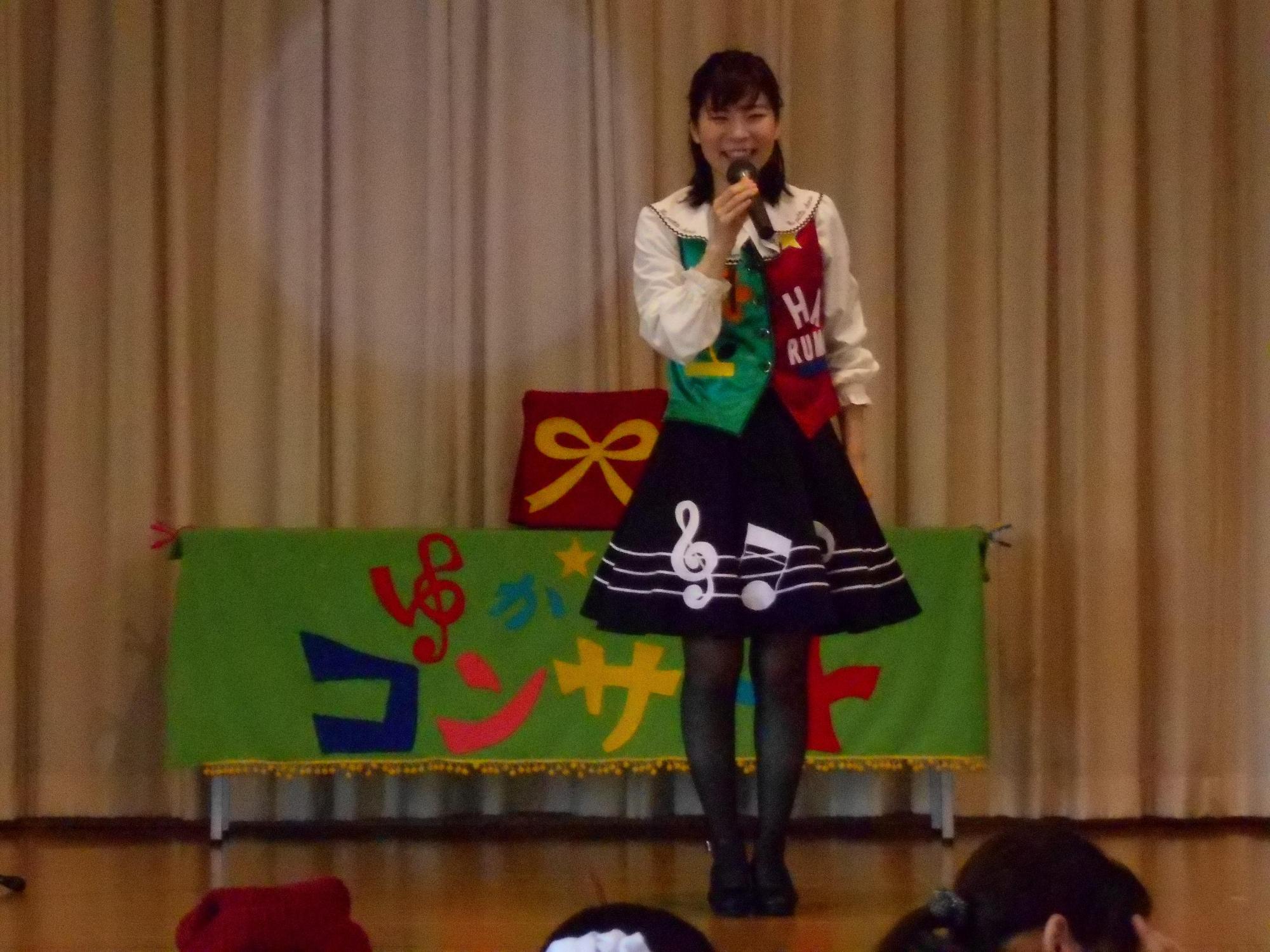ステージの上でスカートをはいた歌のお姉さんがマイクを持ち話をしている写真