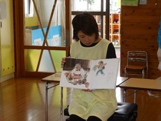 1日保育士体験している保護者が椅子に座り絵本を読んでいる写真