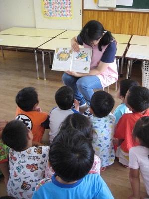椅子に座り絵本の読み聞かせをしている一日保育士体験のお母さんとお話を聞いている園児たちの写真