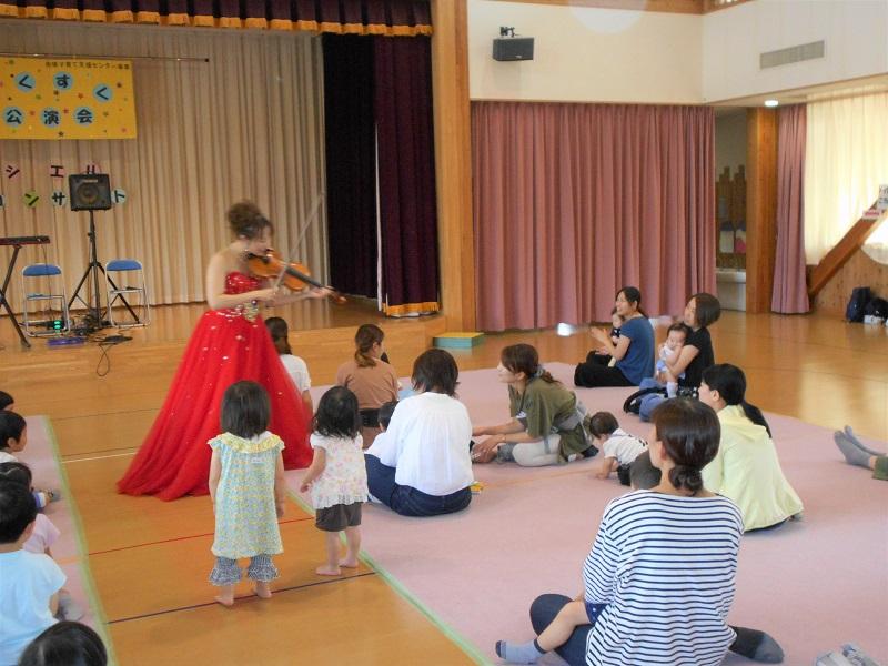 床に座っている参加者親子のすぐ横でバイオリンを演奏する赤いドレスを着た女性の写真