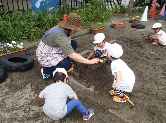 保護者と白い帽子をかぶった園児4人と砂場で遊んでいる写真