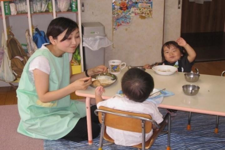 テーブルに座っている2人の園児とその横に床に座って一緒に給食を食べている女性保護者の写真