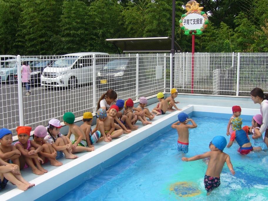 園のプールでプールサイドに座っている園児たちとプールの中で泳いでいる園児たちと一日保育士体験のお母さんの写真