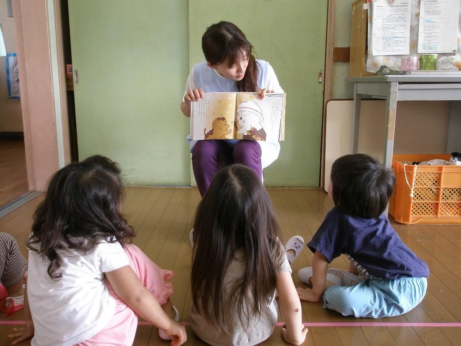 保育園の教室で椅子に座って絵本の読み聞かせをしている紫色のズボンをはいた一日保育士体験のお母さんと読み聞かせを聞いている3人の園児たちの写真