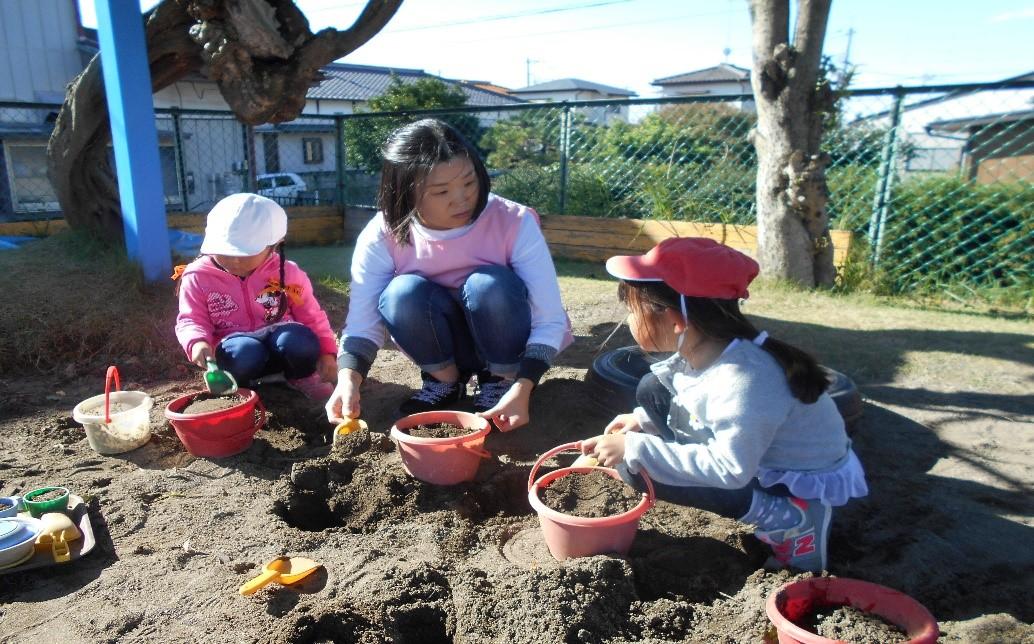 園庭の砂場でバケツに砂を詰めて遊んでいる園児たちと一日保育士体験のお母さんの写真