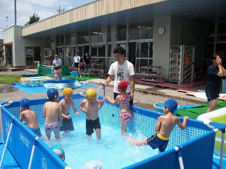 青色の組み立て式のプールに入っている子供たちにホースで水をかけている保護者の写真
