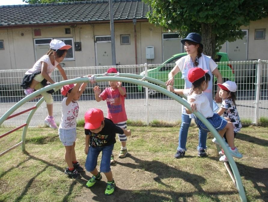 保育園の園庭にある緑色の雲梯で遊んでいる赤白帽子をかぶった6人の園児たちと一日保育士体験のお母さんの写真