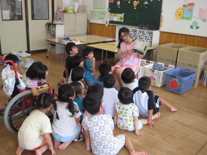 ピンクのエプロンをして絵本の読み聞かせをしている一日保育士体験のお母さんと床に座っている園児たちと車椅子に座っている園児がお話を聞いている写真