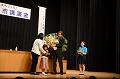 舞台上で女の子より花束を受け取っている二宮清純さんの写真
