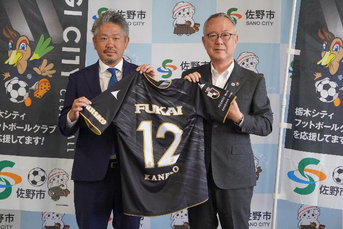 記者会見において栃木シティフットボールクラブの代表取締役社長から佐野市長へユニフォームを手渡ししている写真