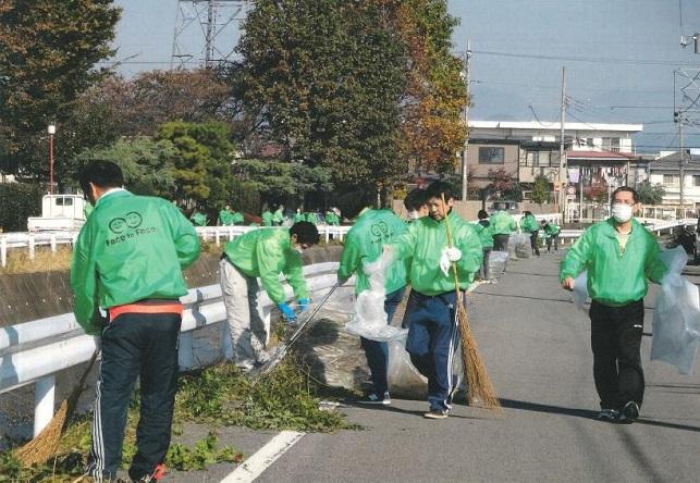 緑の上着を着た信用金庫職員の人々がほうきやごみ袋を持ち川沿いの道路を清掃している写真