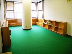 緑のシートが敷かれた床、壁際に本棚があり中央に丸い大きな柱がある児童室の写真
