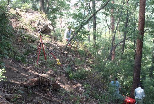 山の斜面で機械を使って地上レーザー測量をしている3名の調査員の写真