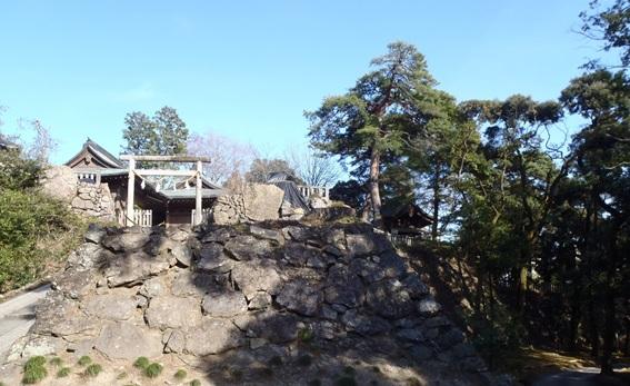 左側は木が生い茂っておりその横には石垣の上に立つ鳥居とその奥に家屋のような建物が建っている写真