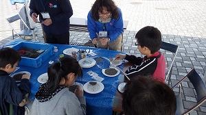 青いシートをひいたテーブルでスタッフと3名の子供が化石探しをしている写真