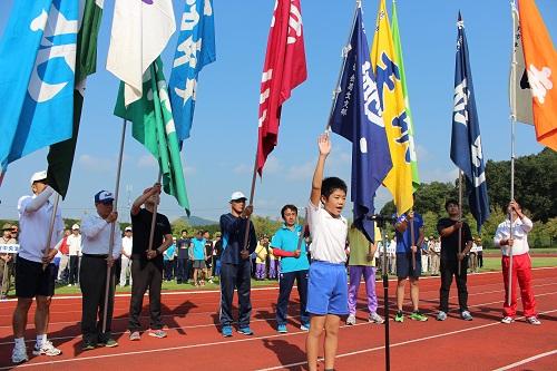 開会式で右手を挙げて選手宣誓をする男の子の後ろで市内14支部の色とりどりの団旗を立てた代表選手が立ち並ぶ写真