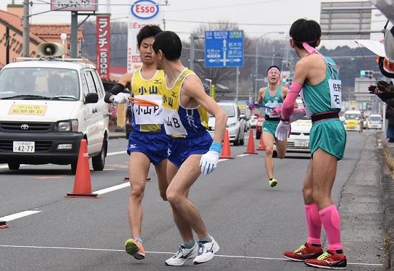 小山市の選手が次の走者に襷を渡している後ろを佐野市の選手が追い着こうと走っている写真