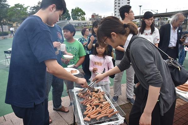 バーベキューコンロで沢山の肉を焼いている男性と女性の横に列を作って待っている人たちの写真