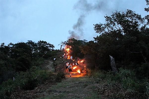 奥では大きな火と黒煙が立ち昇り、参加者が火のついた松明を持ち山道を歩いている様子の写真